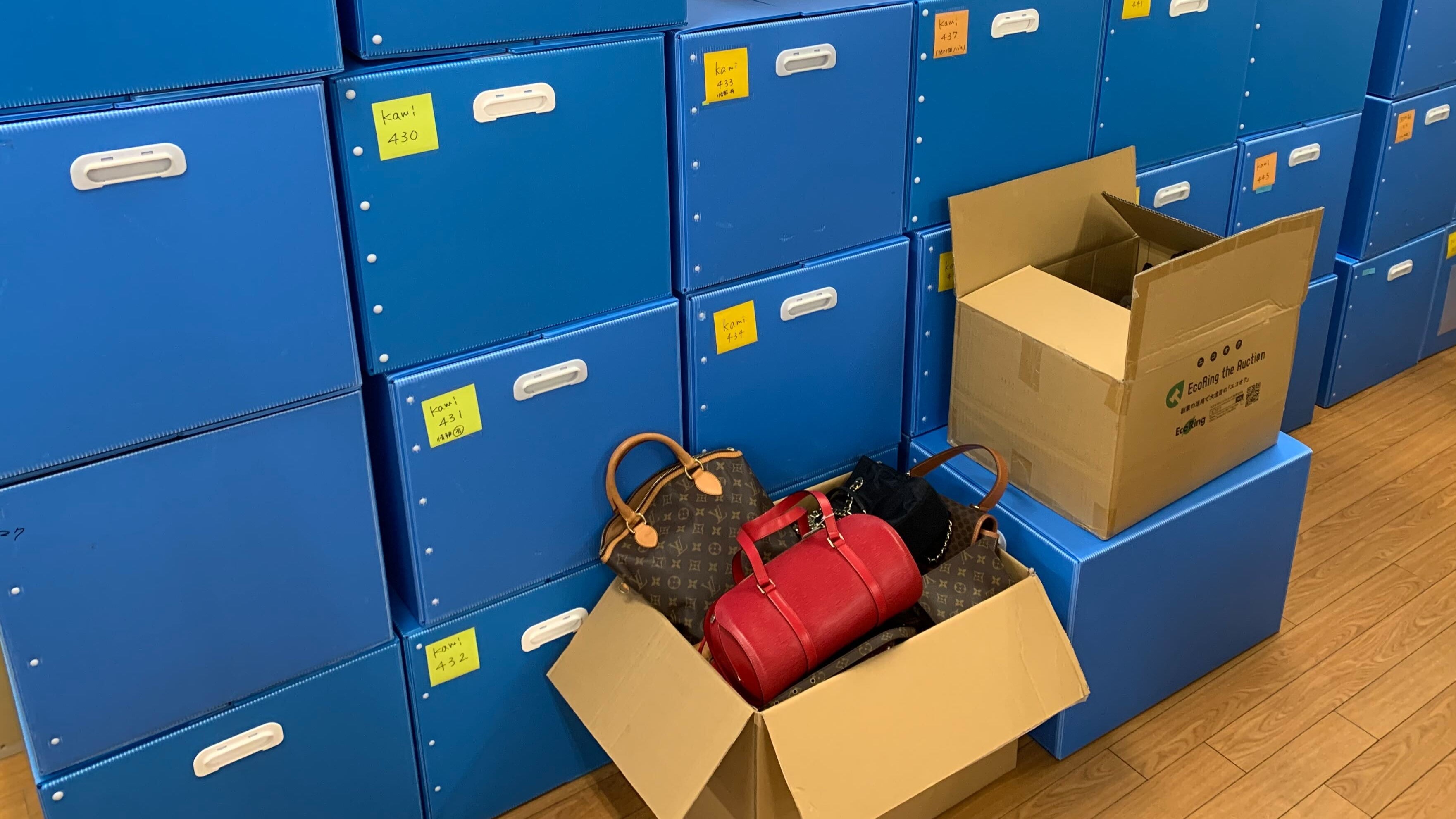 Blue storage boxes with cardboard boxes and a handbag on a wooden floor.